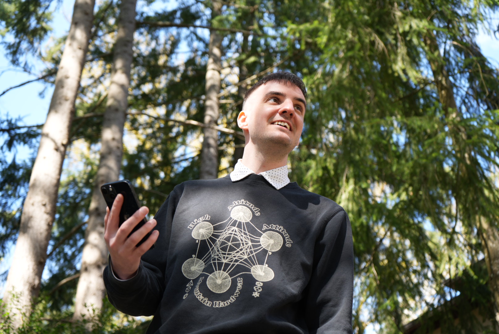 A man outdoors among tall trees, looking up from his phone